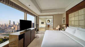 A hotel room with a large bed, a TV on a wooden console, a city view, a bathroom with a mirror, and a nightstand with a lamp.