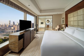 Quarto de hotel com uma cama grande, televisão em um console de madeira, vista para a cidade, banheiro com espelho e mesa de cabeceira com abajur.