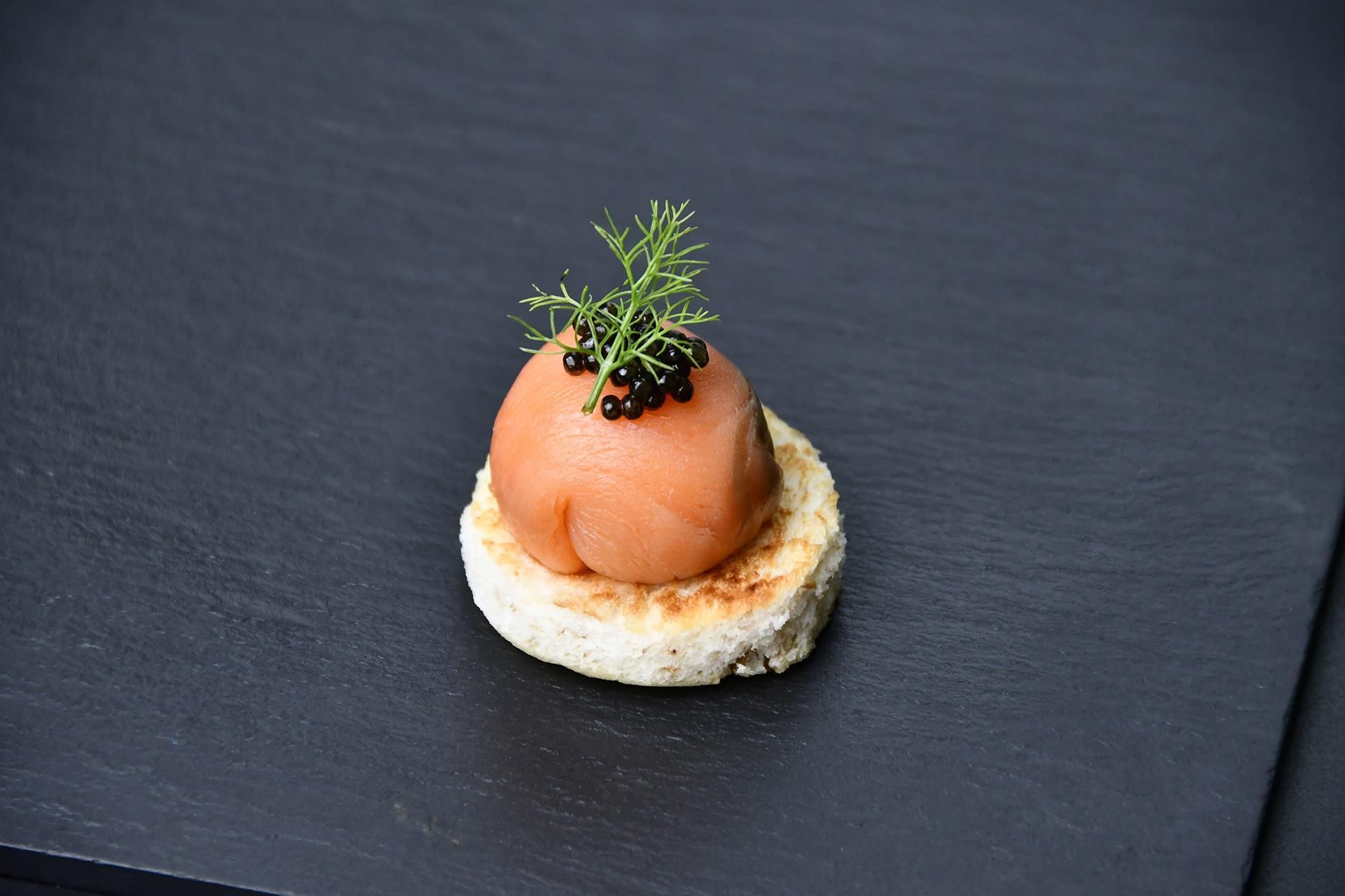 A round, salmon-based appetizer on a piece of bread, garnished with herbs and black caviar, presented on a dark slate surface.
