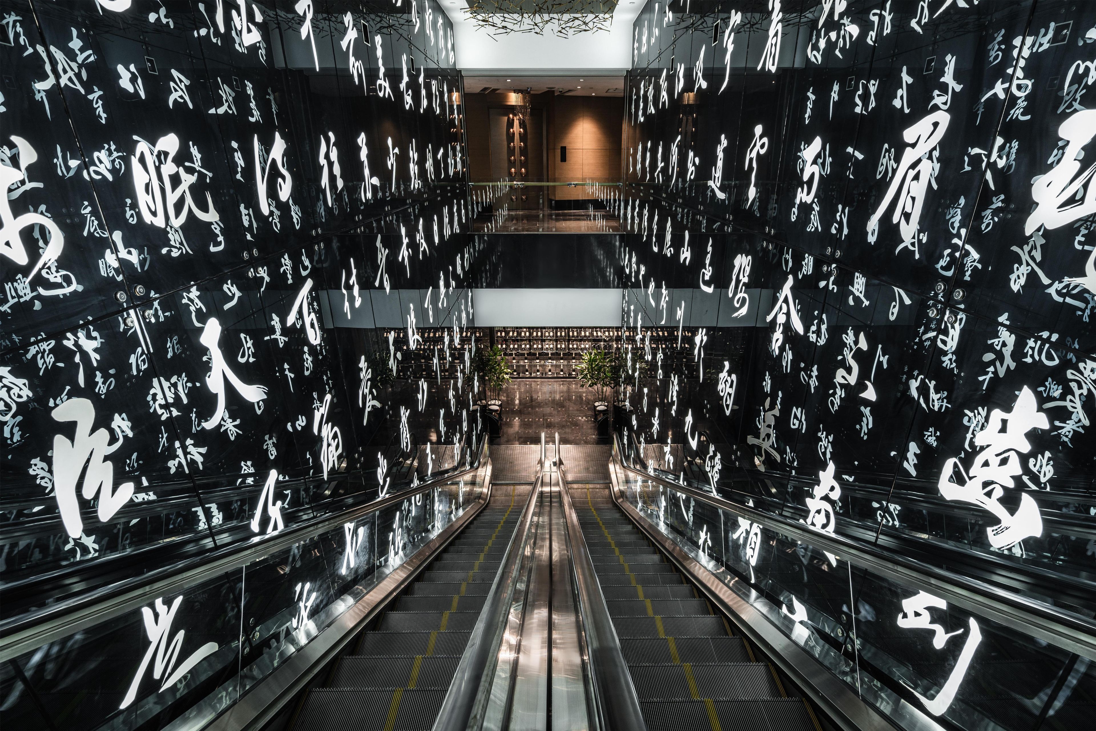 Un pasillo interior con escaleras mecánicas, flanqueado por paredes cubiertas de caligrafía blanca.