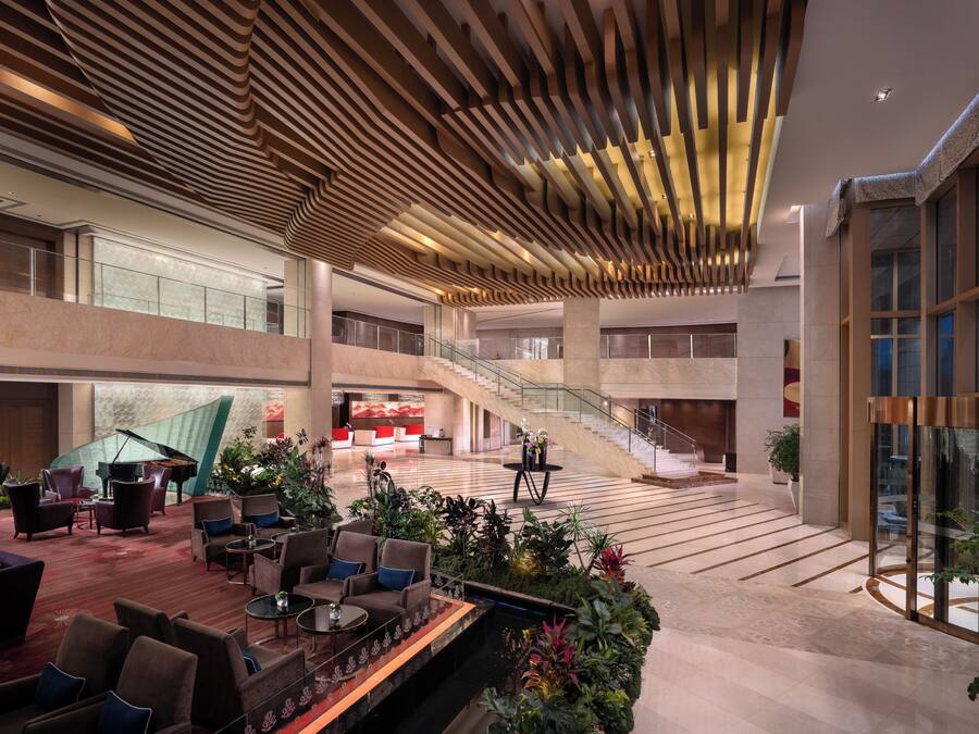 A spacious lobby with a grand piano, multiple seating areas, a staircase, and a large glass wall.