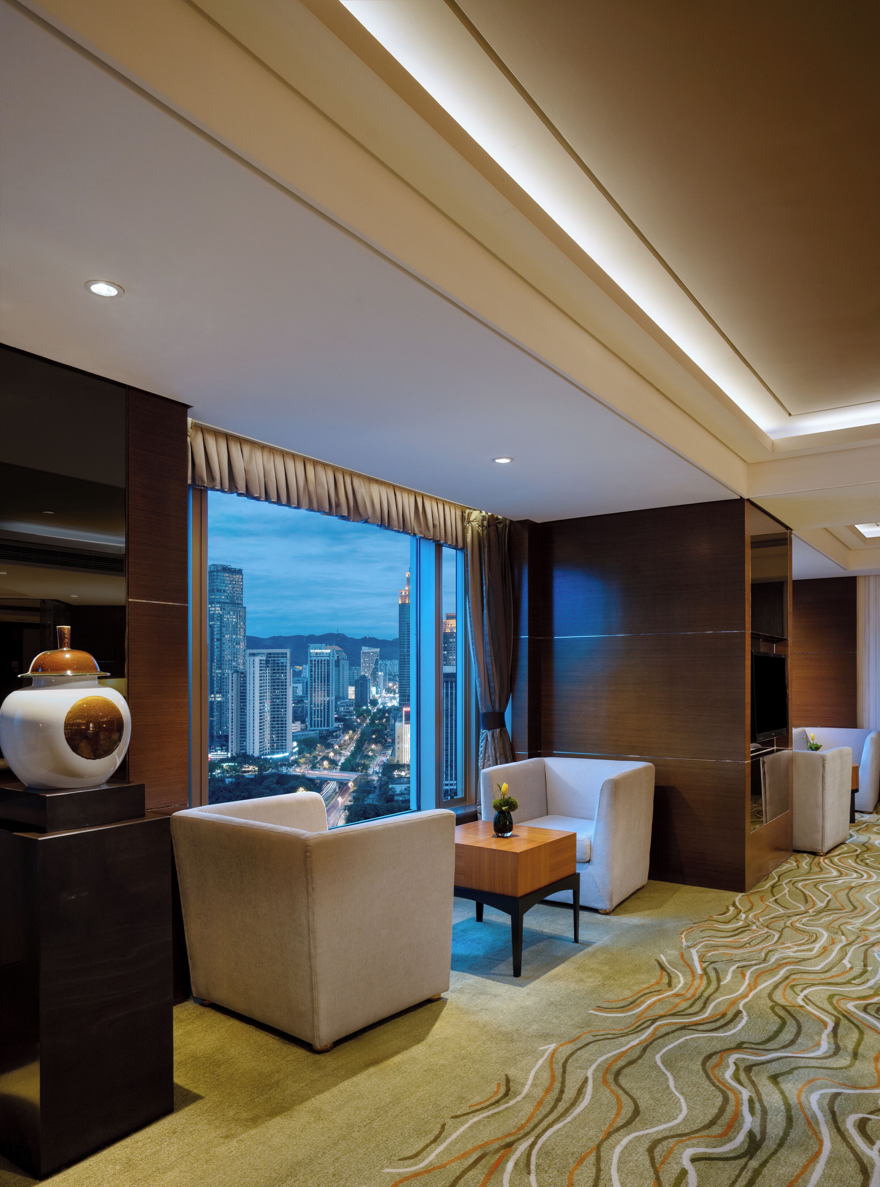 Habitación de hotel con un ventanal grande que ofrece vistas a la ciudad, un sofá, una mesita pequeña y un jarrón decorativo.