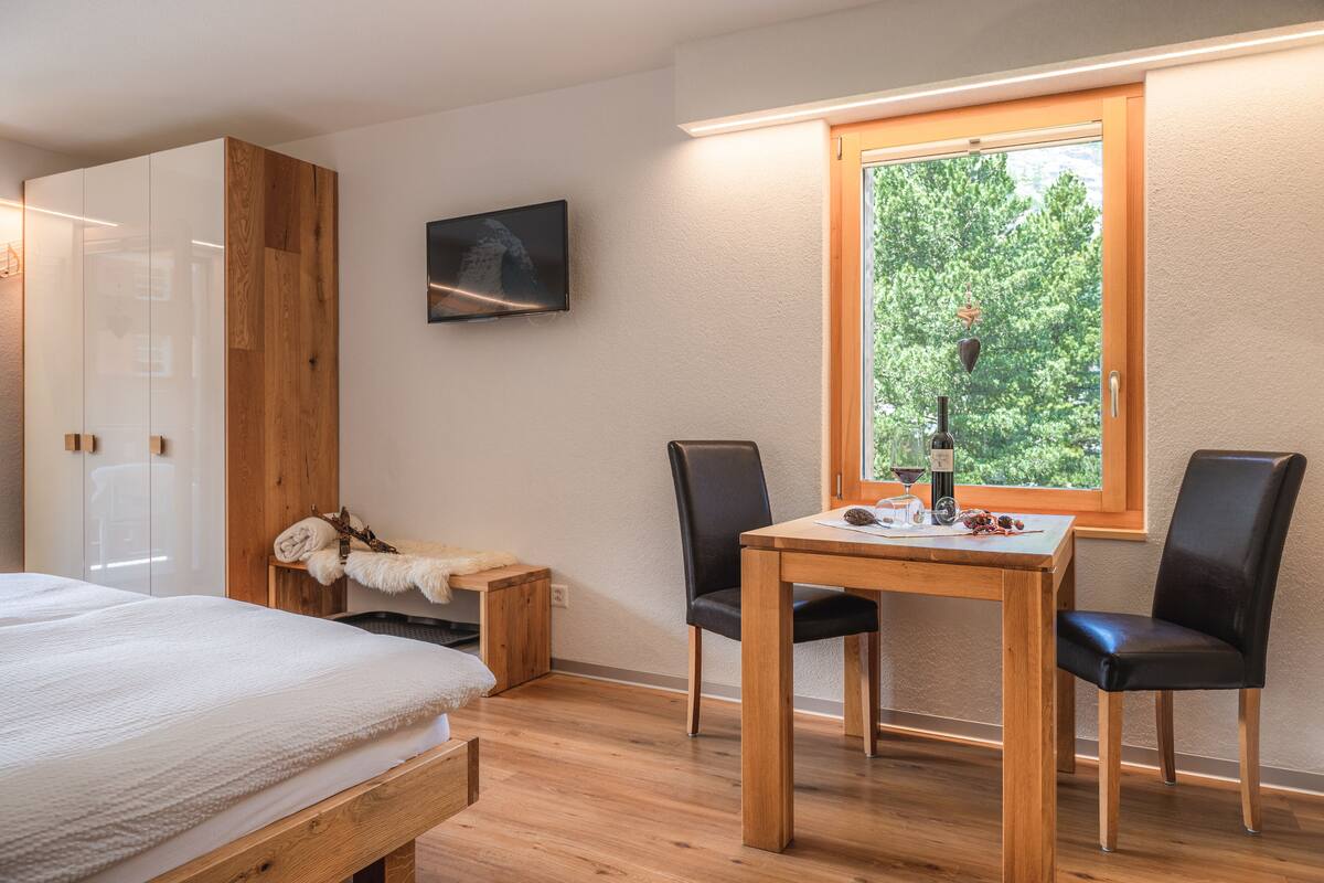 Un dormitorio con cama, armario de madera, una mesita con botella y vasos, y ventana con vista a árboles.