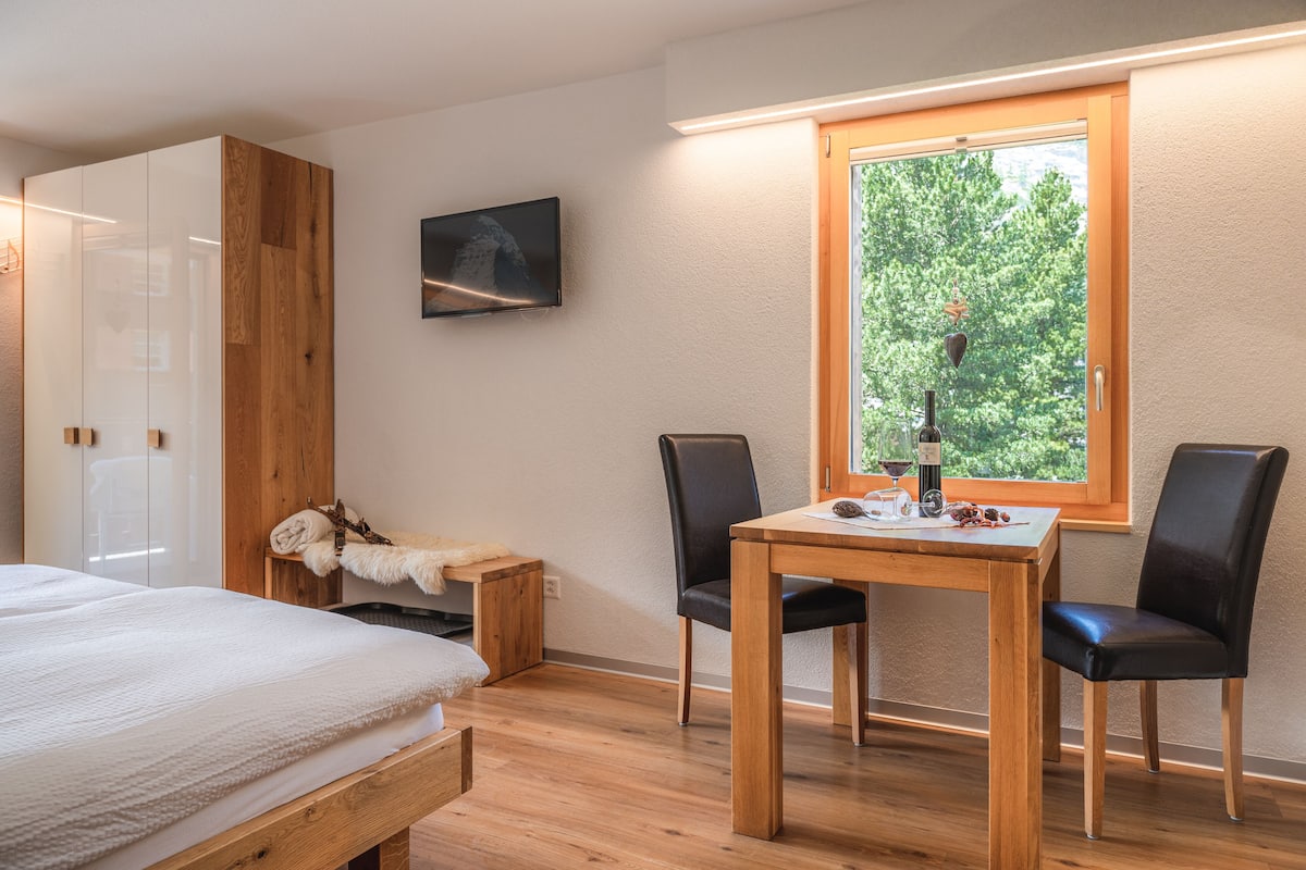 A bedroom with a bed, a wooden wardrobe, a small table with a bottle and glasses, and a window with a view of trees.