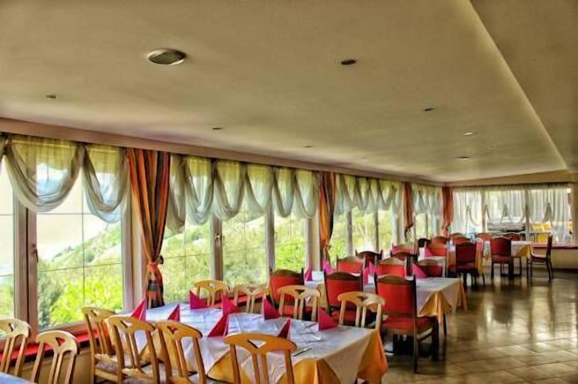 Un espace repas avec des tables et des chaises, de grandes fenêtres avec des rideaux et une vue sur la verdure à l’extérieur.