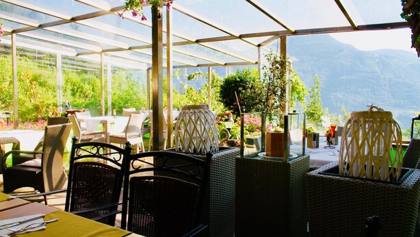 Un restaurante de estilo invernadero con mobiliario de mimbre, plantas en macetas y vistas a montañas.