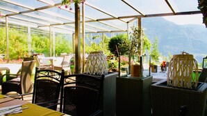 A greenhouse-style restaurant with wicker furniture, potted plants, and a view of mountains. - Hotel Unterinnerhof (Renon)