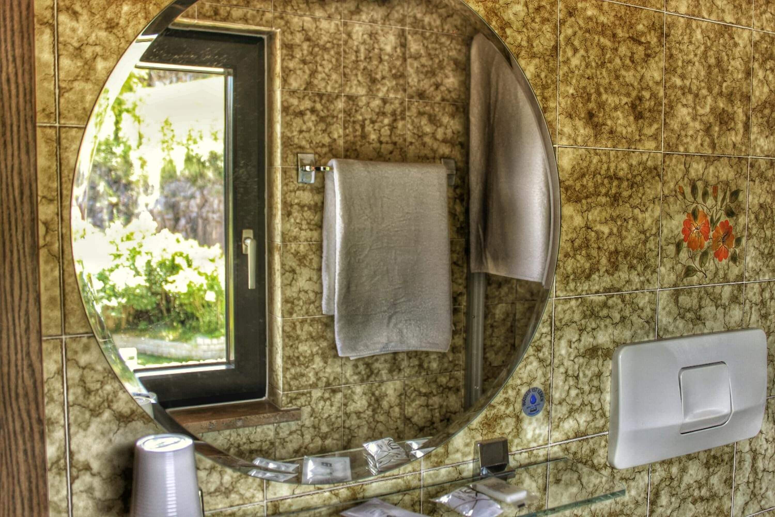 Een badkamer met een ronde spiegel, een handdoekhouder, een wastafel en een raam met uitzicht op groen.