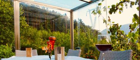 A table with a pizza and a glass of red wine, set in a glass-enclosed patio with wicker furniture and a view of greenery and hills.