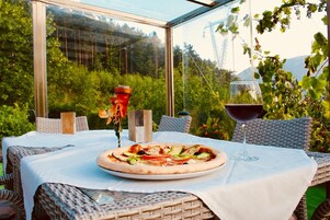 Una mesa con pizza y una copa de vino tinto, ubicada en un patio cubierto de vidrio con mobiliario de mimbre y vistas a la vegetación y colinas.