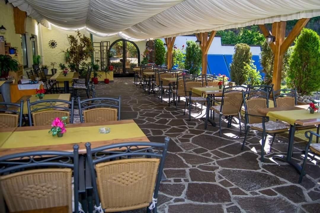 Een terras buiten met stenen vloer, rieten tafels en stoelen, omringd door groen en een blauwe muur.