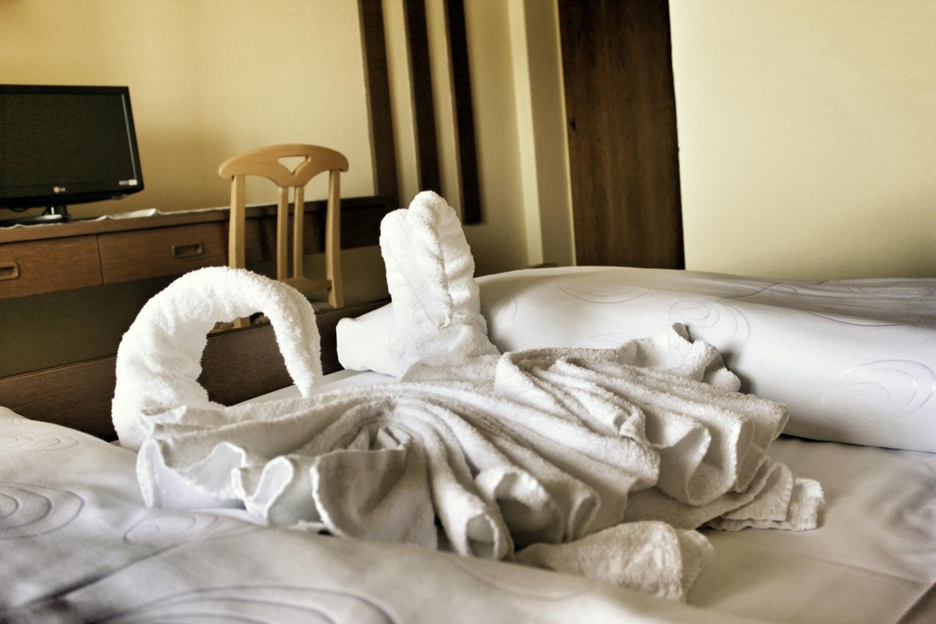 Ein Hotelzimmer mit einem Bett, einem Holzstuhl, einem Schreibtisch mit Computerbildschirm und einer Wand mit vertikalen Jalousien.