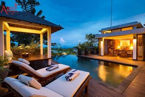 Un espace extérieur luxueux avec une piscine, des chaises longues, un coin salon abrité et un bâtiment moderne à l’éclairage chaleureux.