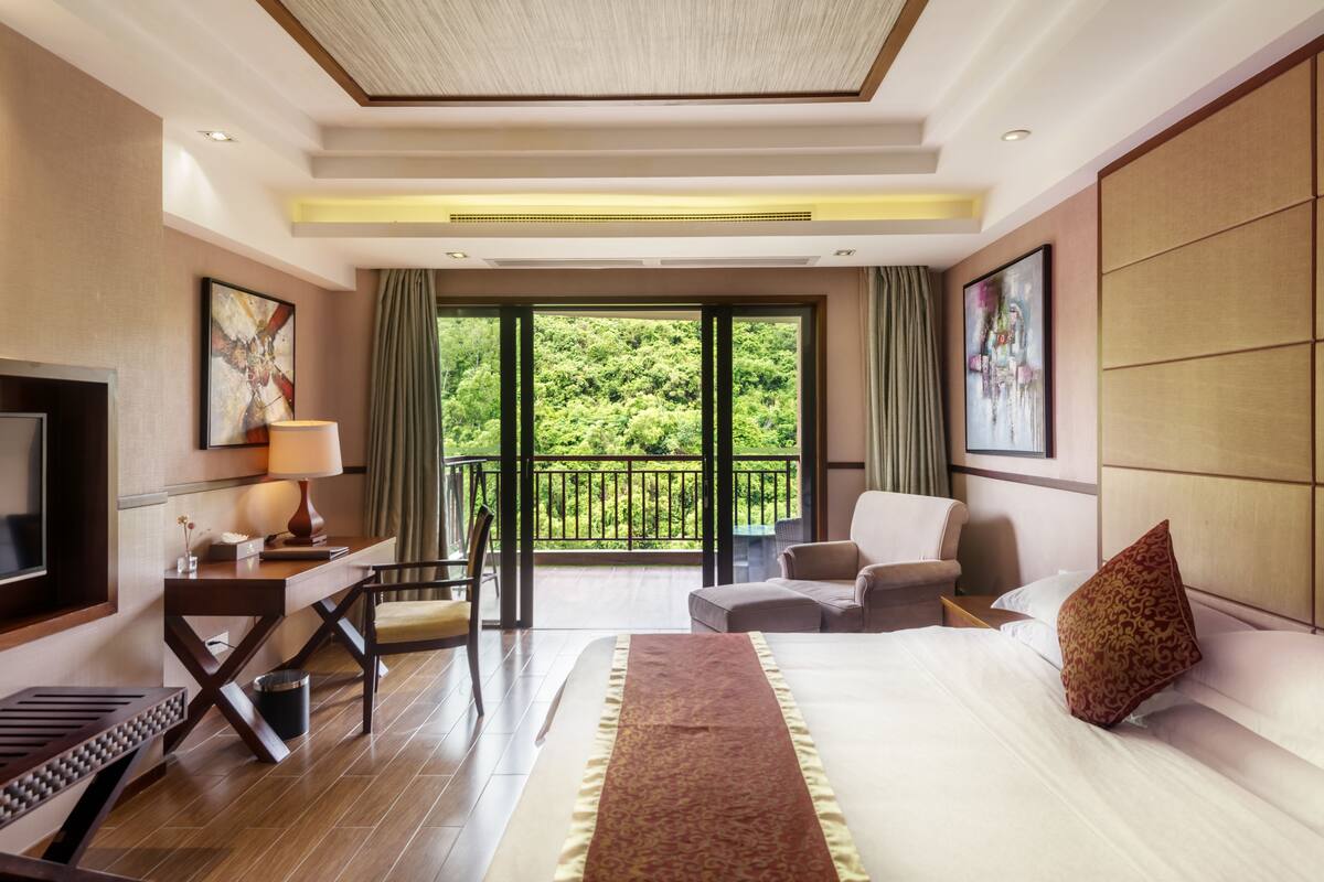 Une chambre d’hôtel avec un grand lit, un bureau avec une lampe, une télévision, un balcon donnant sur un espace verdoyant et un canapé.