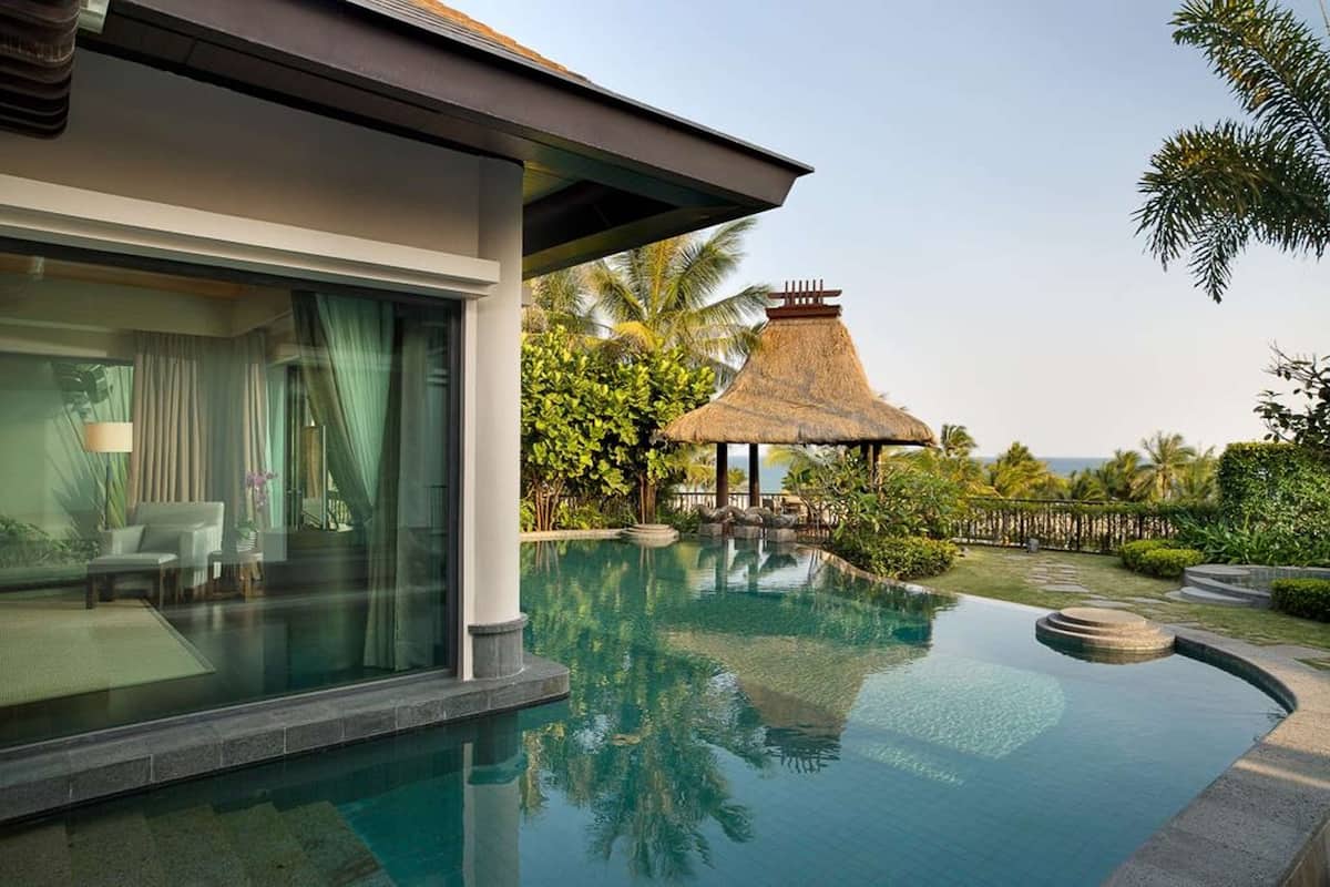 A modern villa with a glass-enclosed pool, a thatched-roof gazebo, and lush tropical landscaping.