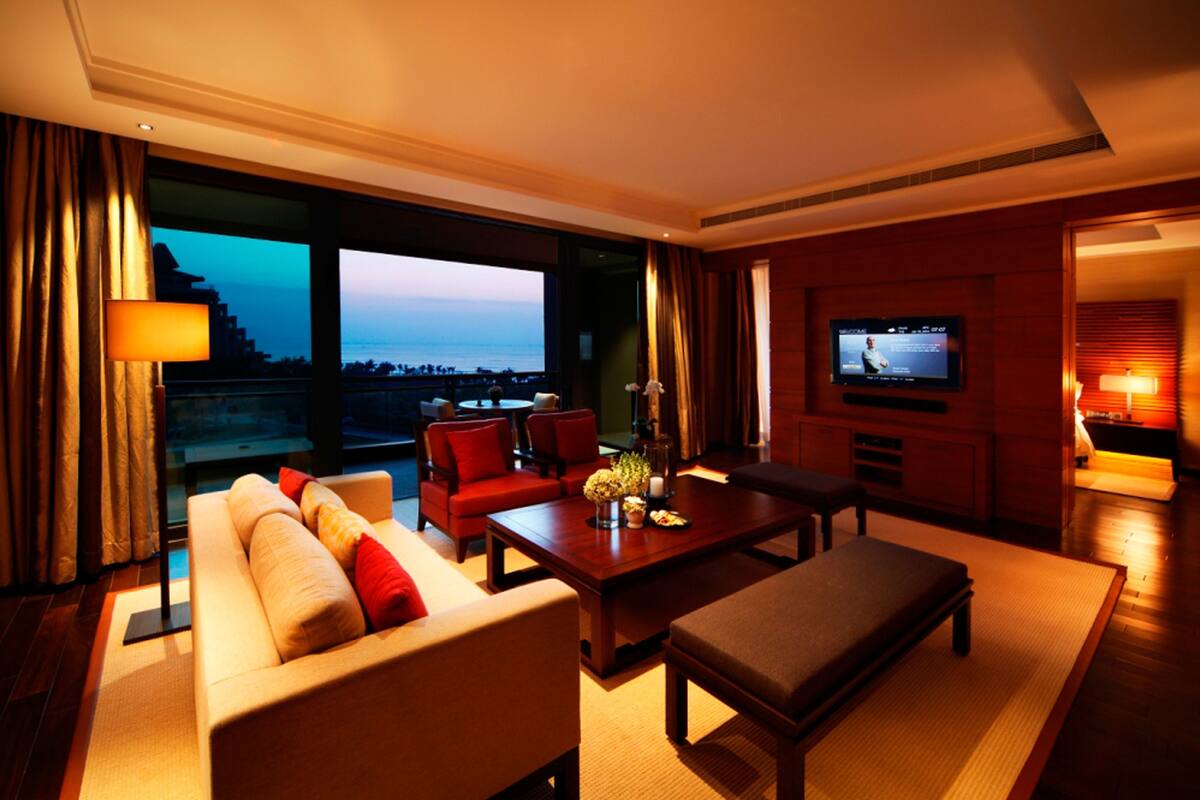 A modern hotel room with a large window offering a view of the sea, a comfortable seating area with a sofa and chairs, a flat-screen TV mounted on the wall, and a small table with a vase of flowers.