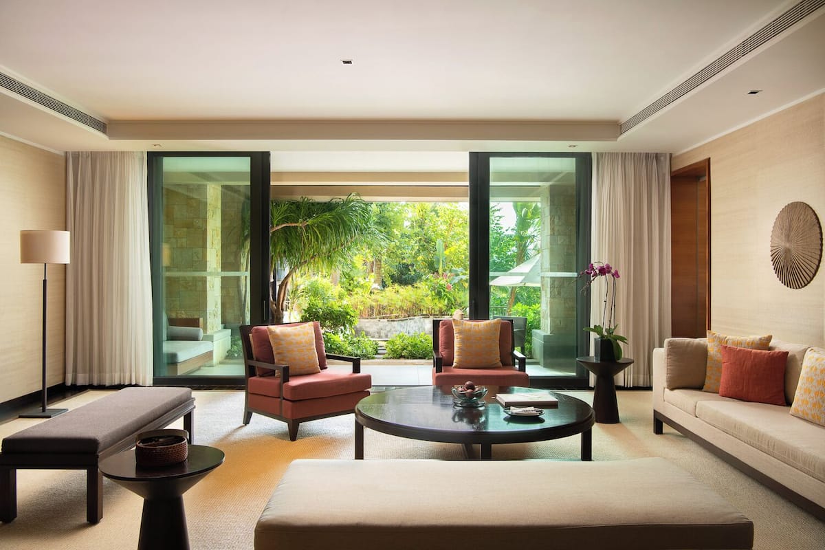 A modern hotel room with a sofa, two armchairs, a coffee table, and a large sliding glass door leading to a garden.