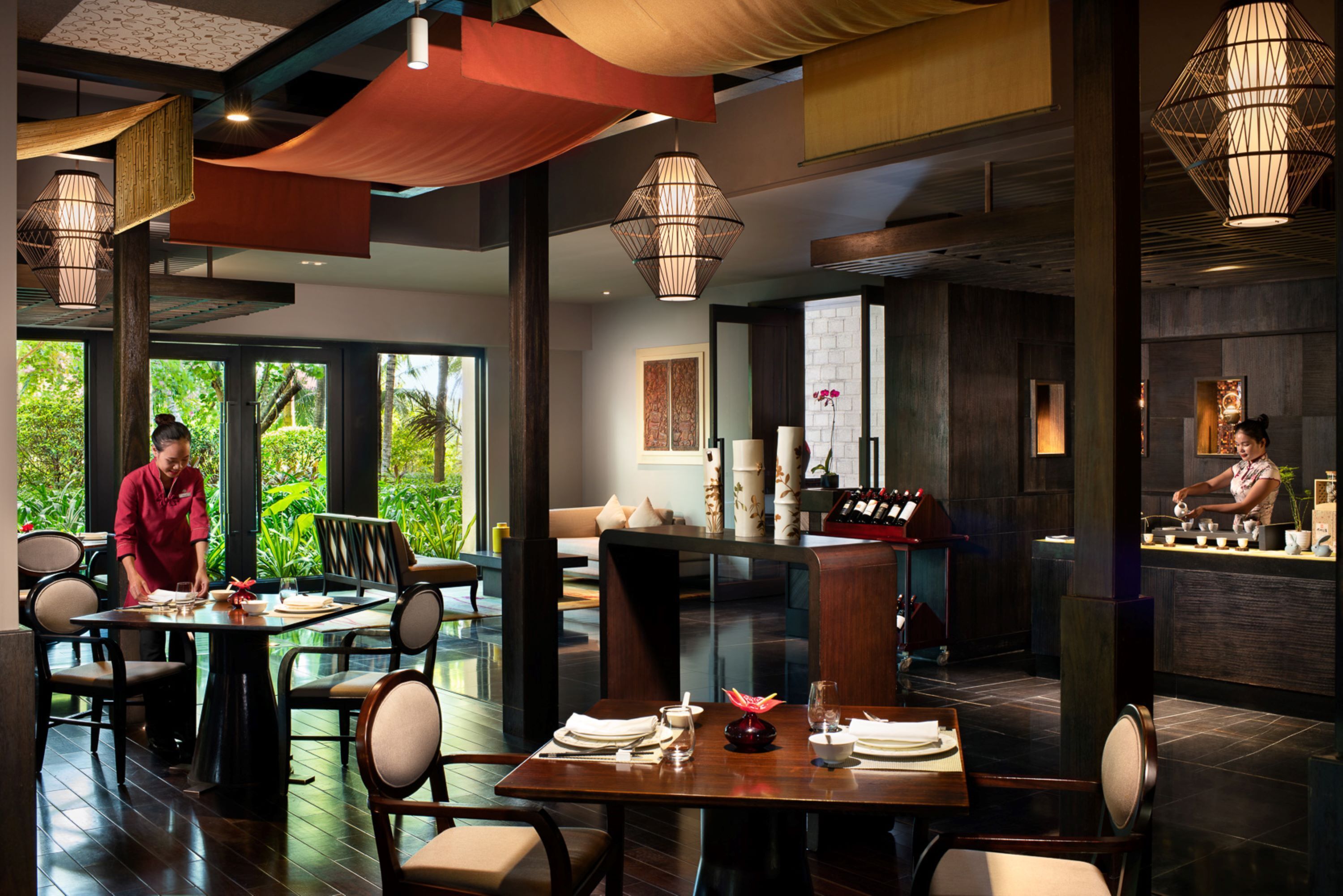 Un restaurant avec des tables dressées pour le service, un coin bar et une vue sur la verdure environnante.