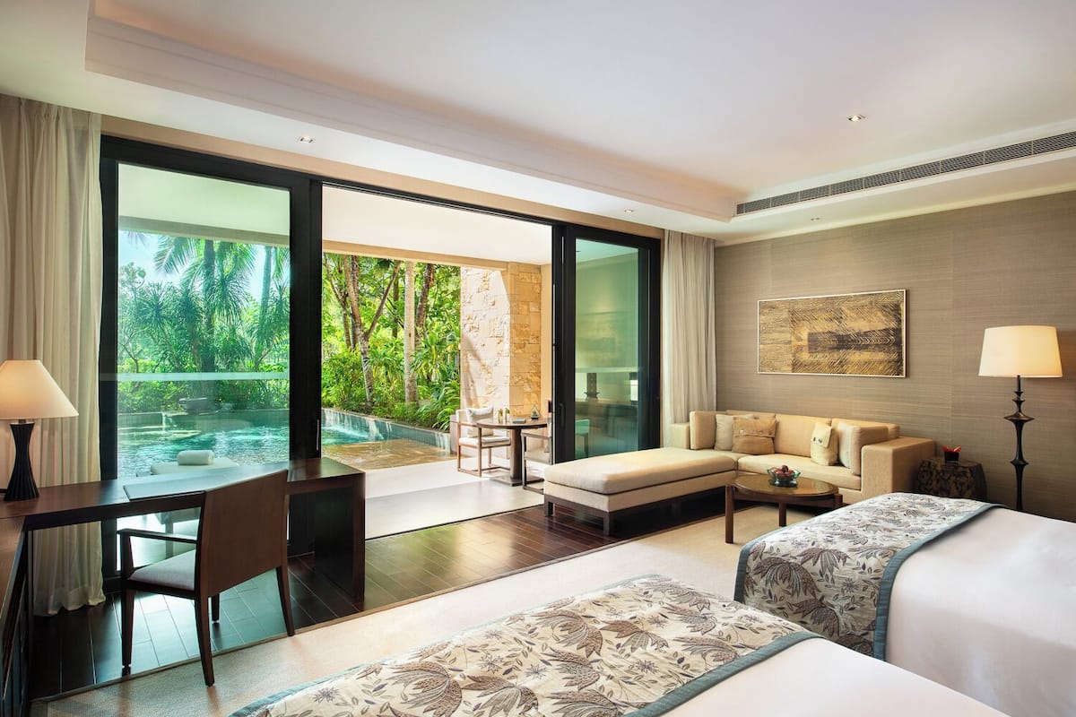 A hotel room with a large sliding glass door to a pool, a sofa, a bed, a desk, and a lamp.