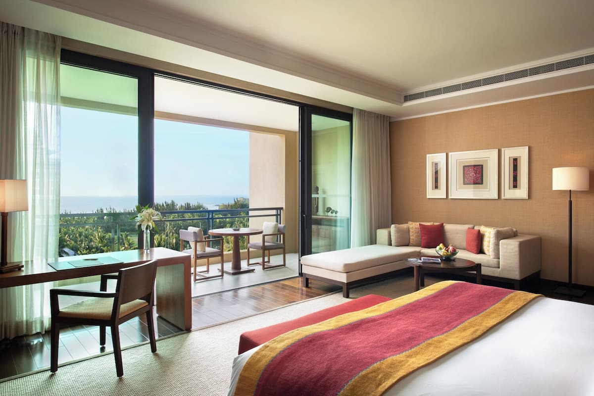 A hotel room with a bed, a desk, a chair, a sofa, a small table, and a view of the ocean.