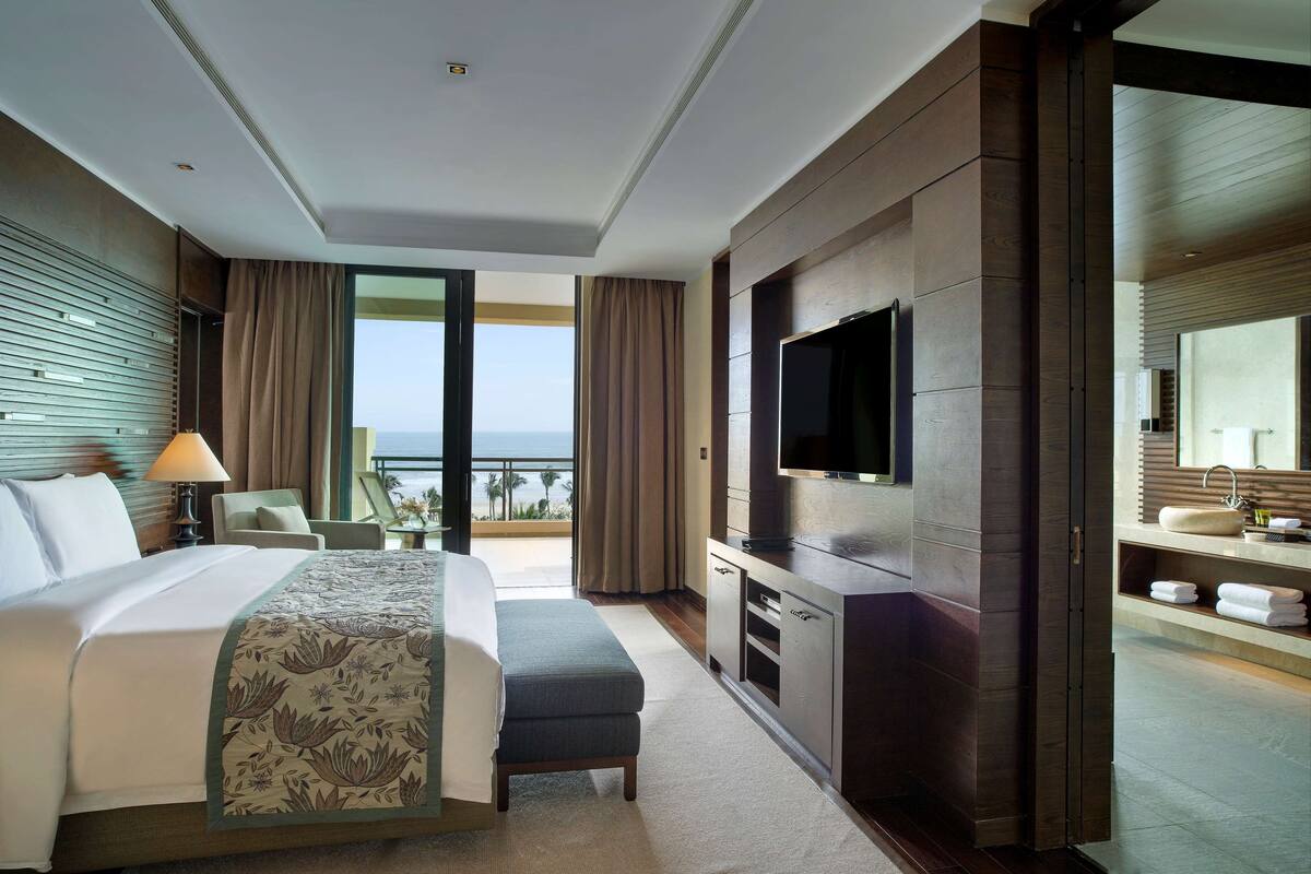 A modern hotel room with a large bed, a TV, a desk, and a balcony with a view of the ocean.