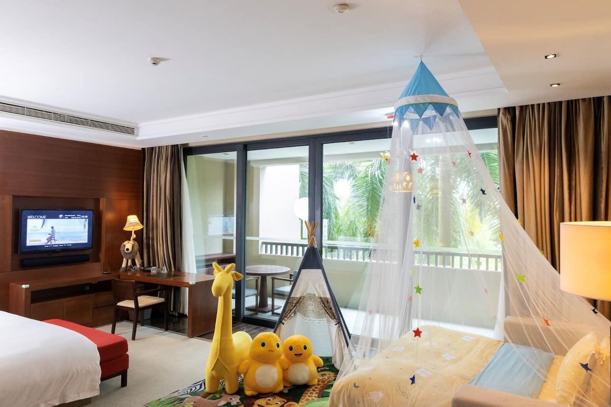 A hotel room with a bed, a desk, a television, a chair, a giraffe and duck plush toys, a tent, and a balcony with curtains.