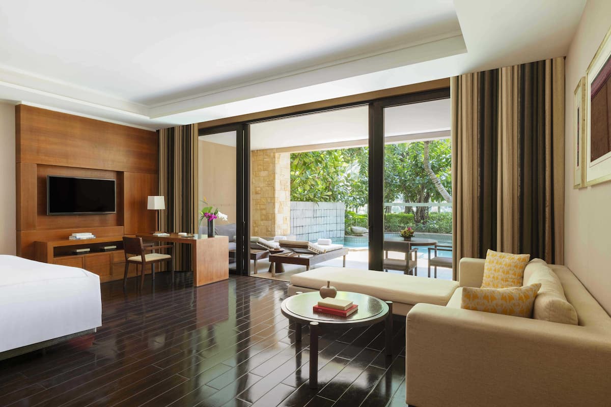A hotel room with a bed, a TV, a desk, a sofa, and a view of a pool and greenery.