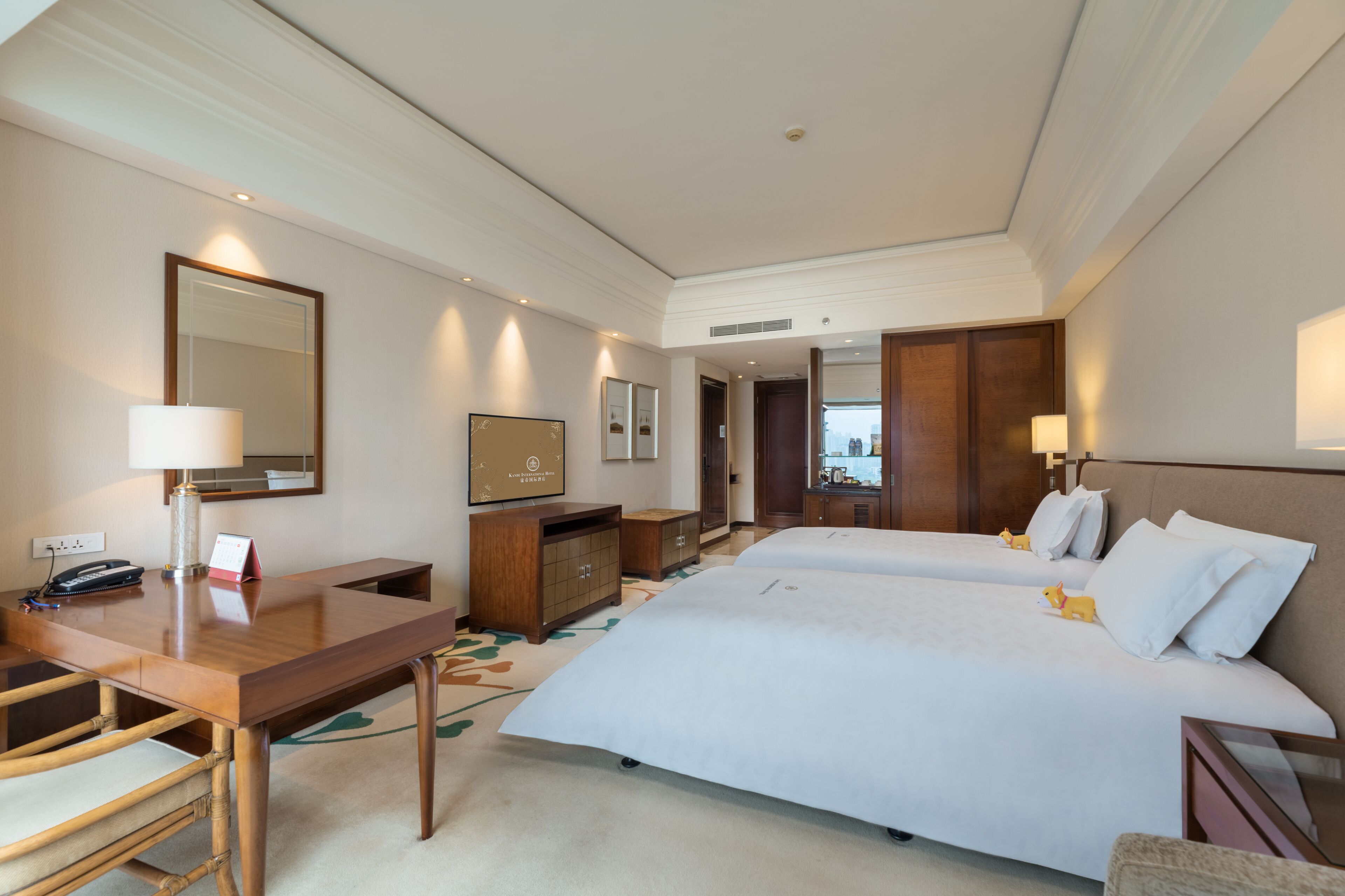 A hotel room with a wooden desk, two beds, a TV, and a mirror.