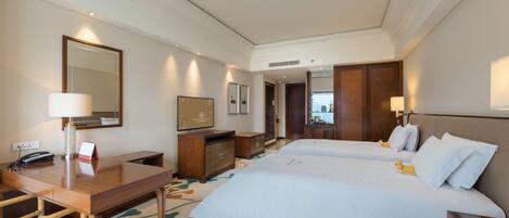 A hotel room with a wooden desk, two beds, a TV, and a mirror.