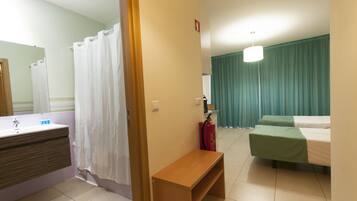Habitación de hotel con baño, un banco de madera, cama con cubrecama verde y mesita de noche con extintor rojo.