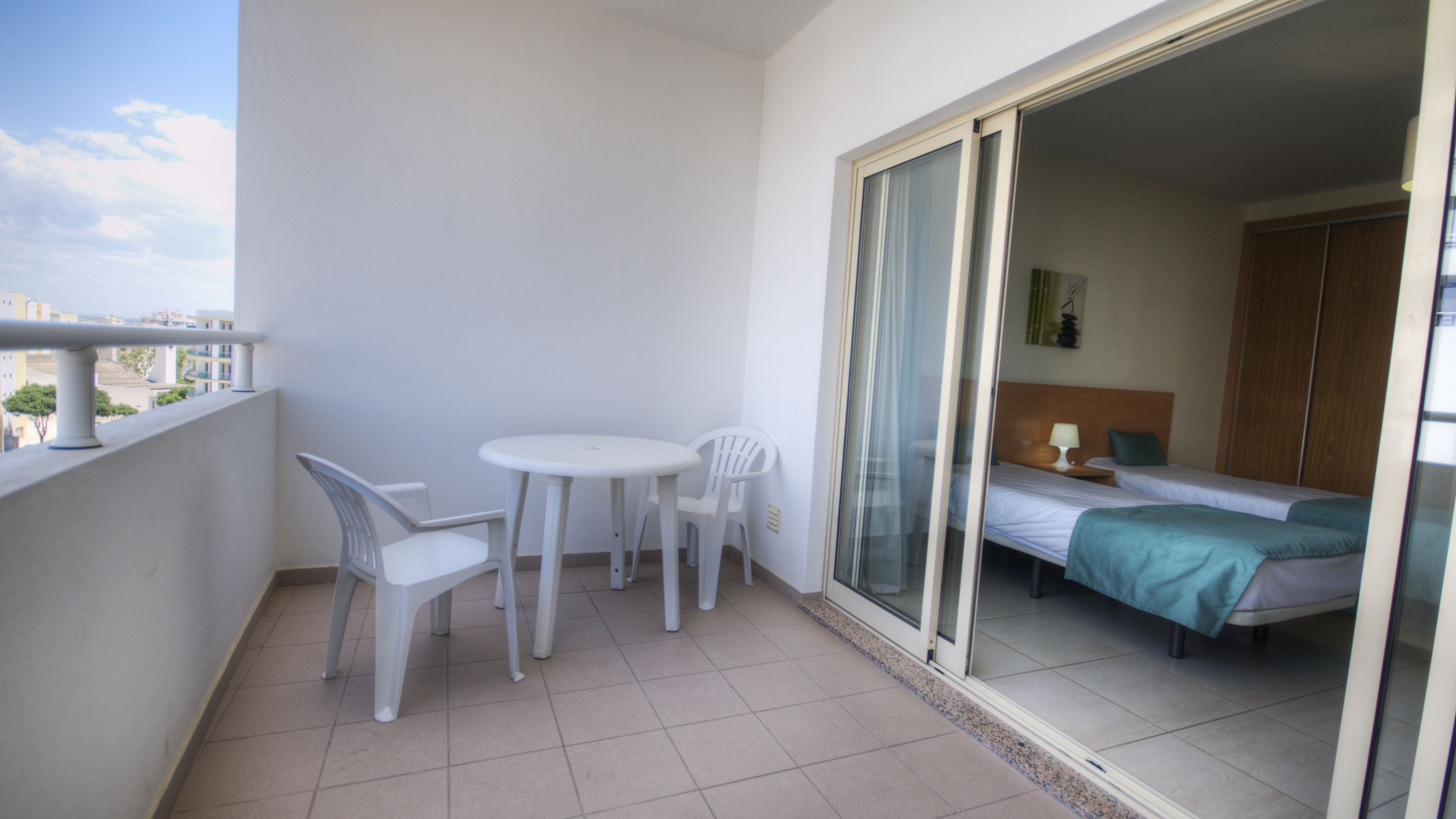 A balcony with a table and chairs, a sliding glass door leading to a bedroom with a bed and a nightstand.
