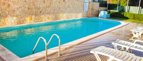 A swimming pool with a stone wall, tiled deck, and lounge chairs.