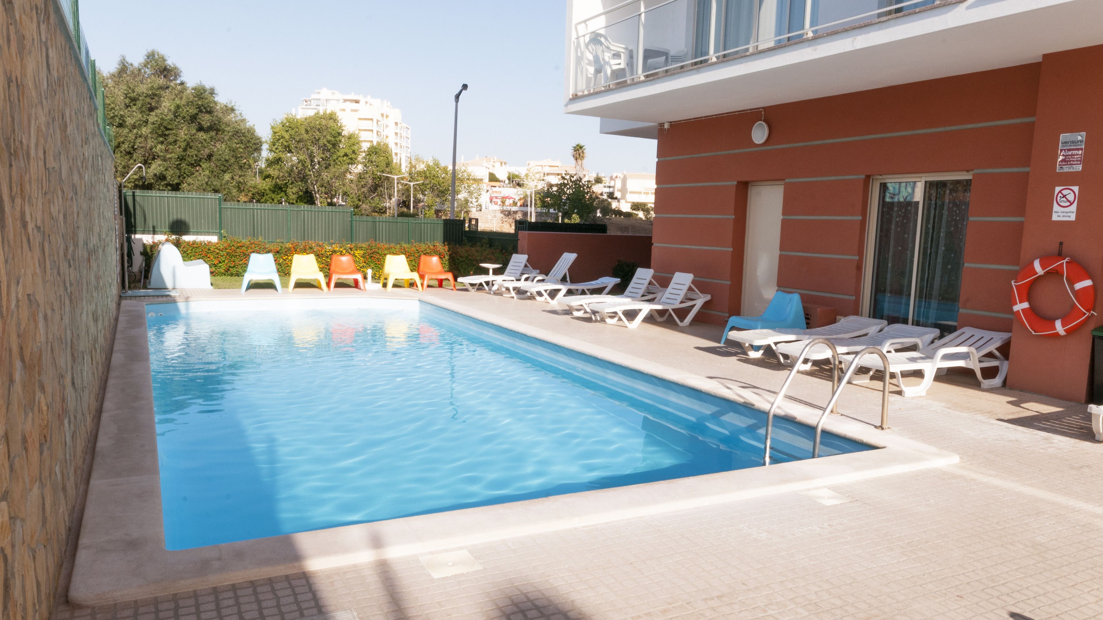 Une piscine dotée de chaises longues et d’une bouée de sauvetage, entourée d’un bâtiment à la façade rouge et doté d’un balcon.