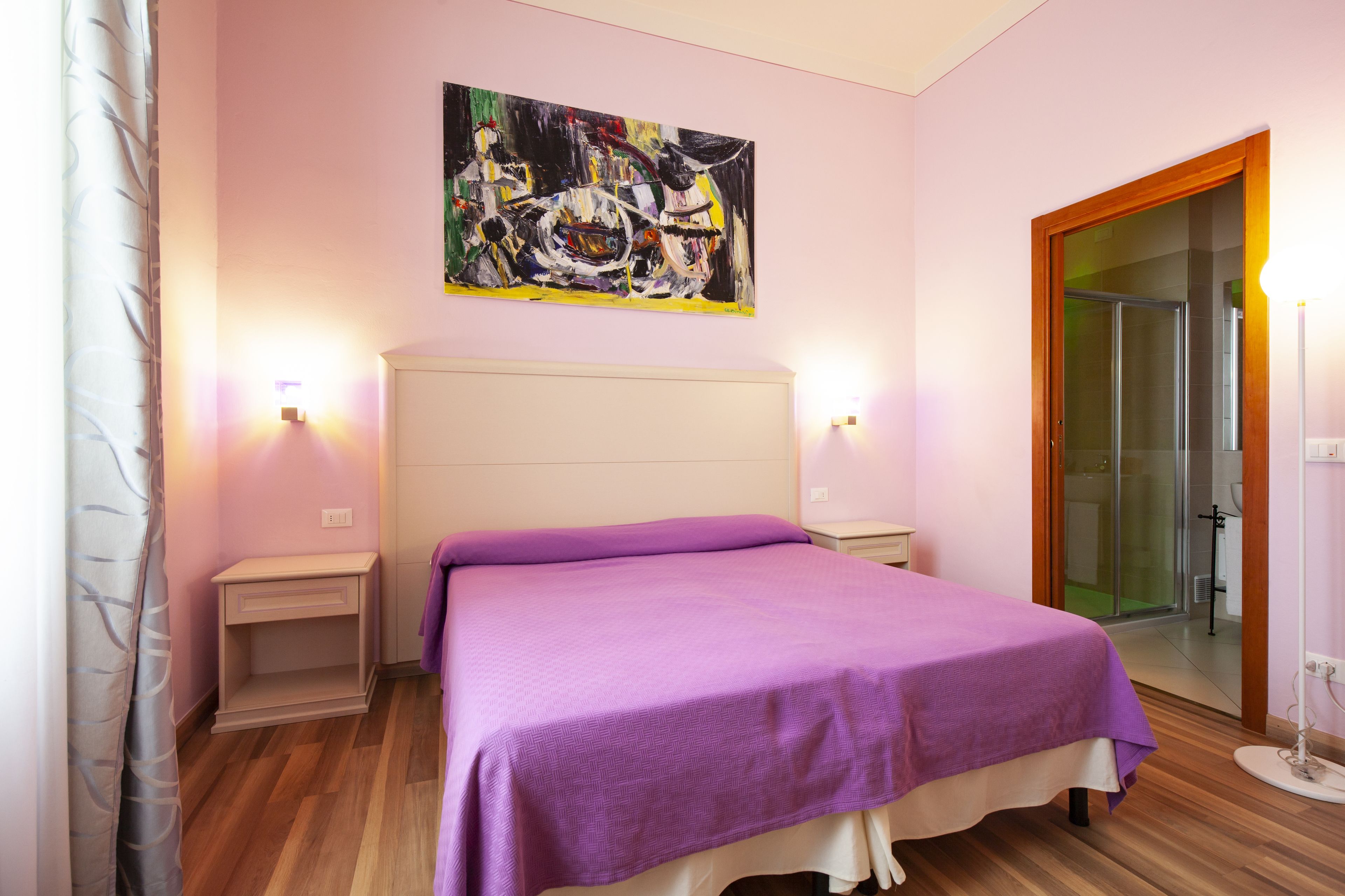 A bedroom with a bed, a nightstand, a bedside table, a painting on the wall, and a door leading to a bathroom.