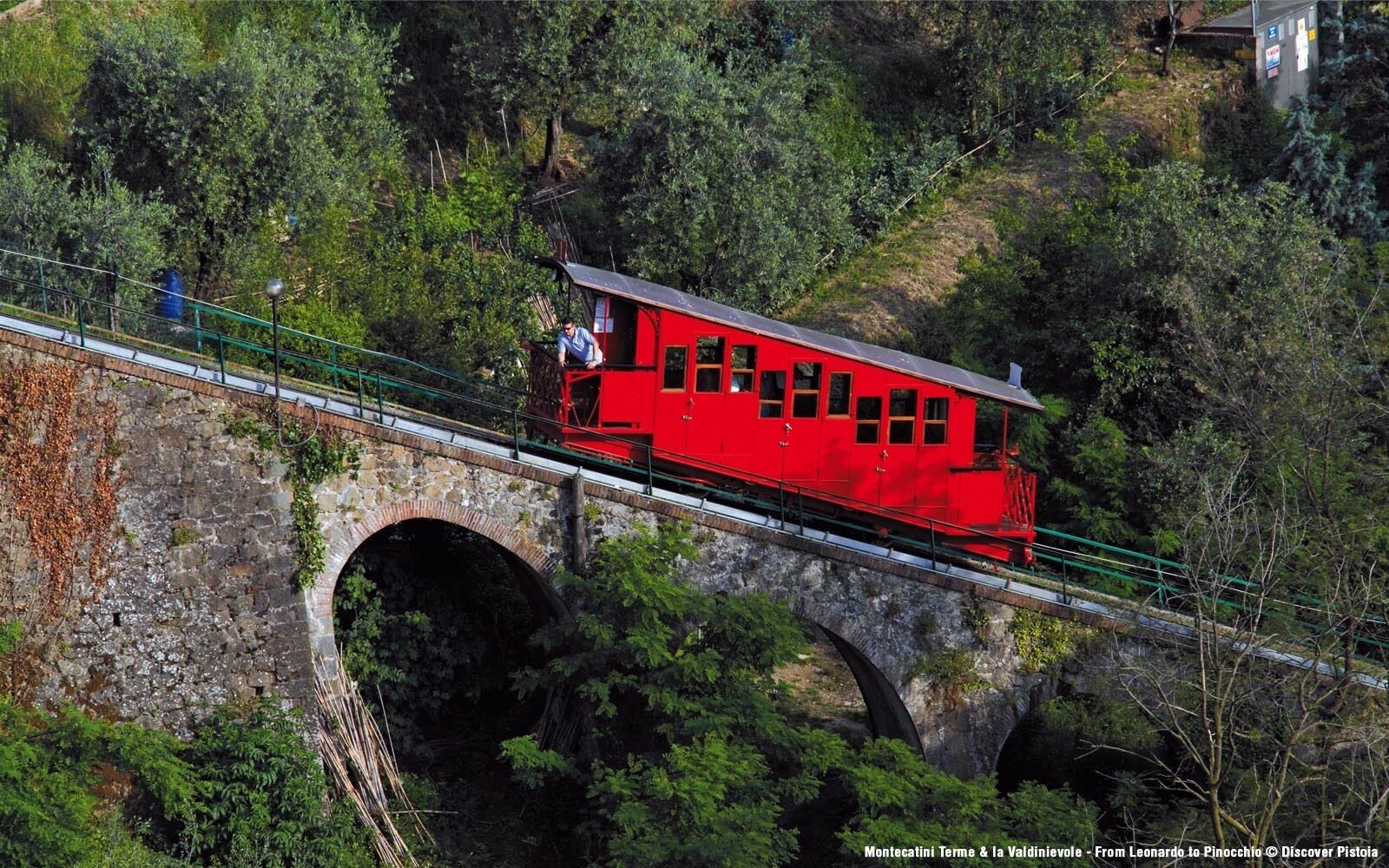 Un teleférico rojo sobre un puente de piedra con vegetación a su alrededor.