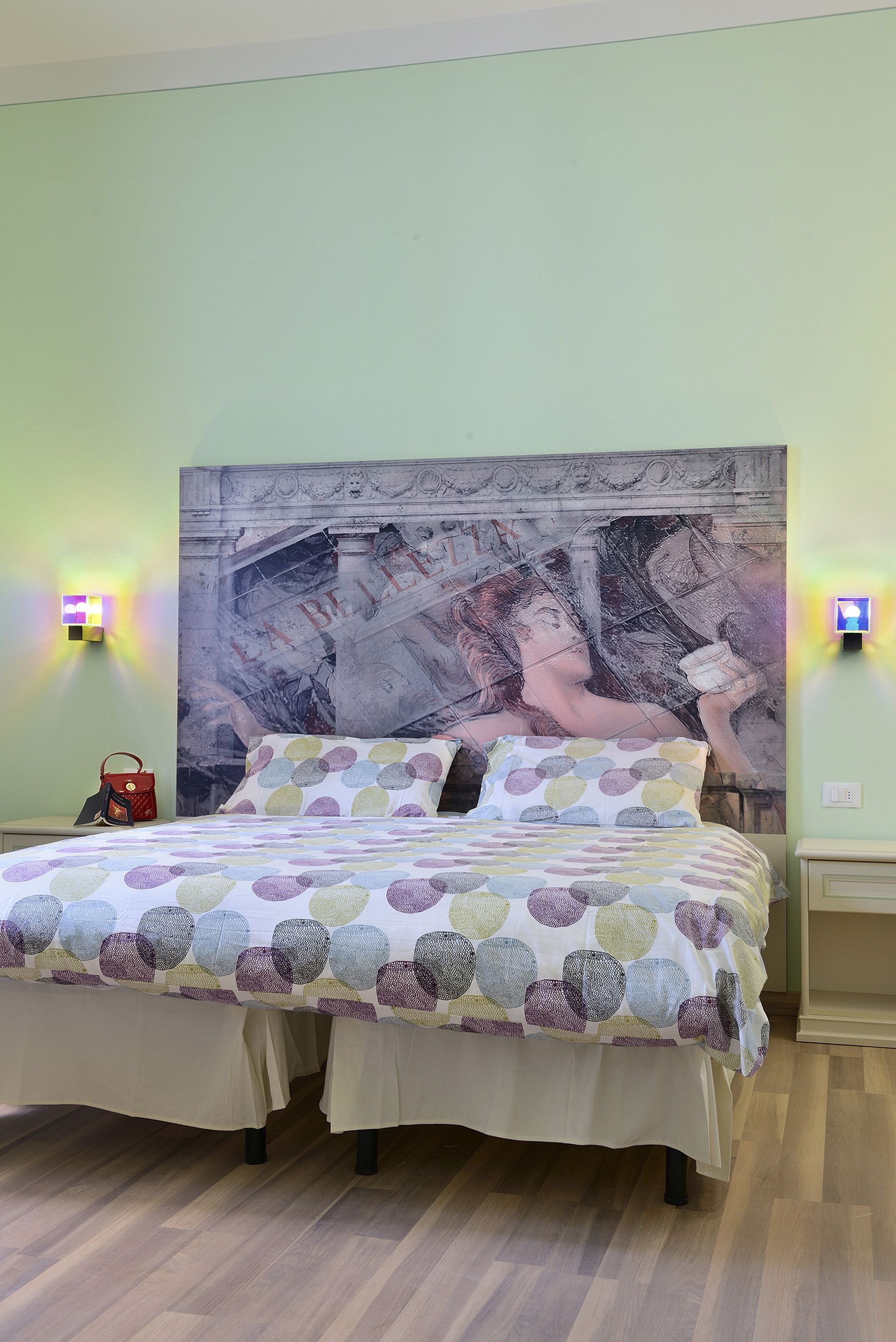 A bedroom with a large bed, a headboard with a mural, two wall-mounted lights, and a nightstand with a red bag.