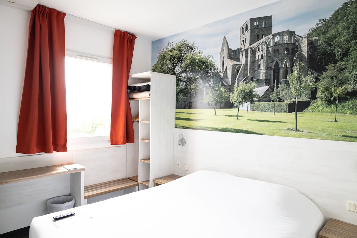A bedroom with a large bed, a window with red curtains, a wardrobe, and a mural of a castle on the wall.