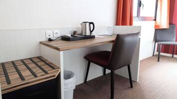 A compact hotel room with a wooden desk, a chair, a coffee maker, and a trash bin.