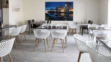 A modern dining area with white lattice chairs and dark tables, featuring a large cityscape painting on the back wall.