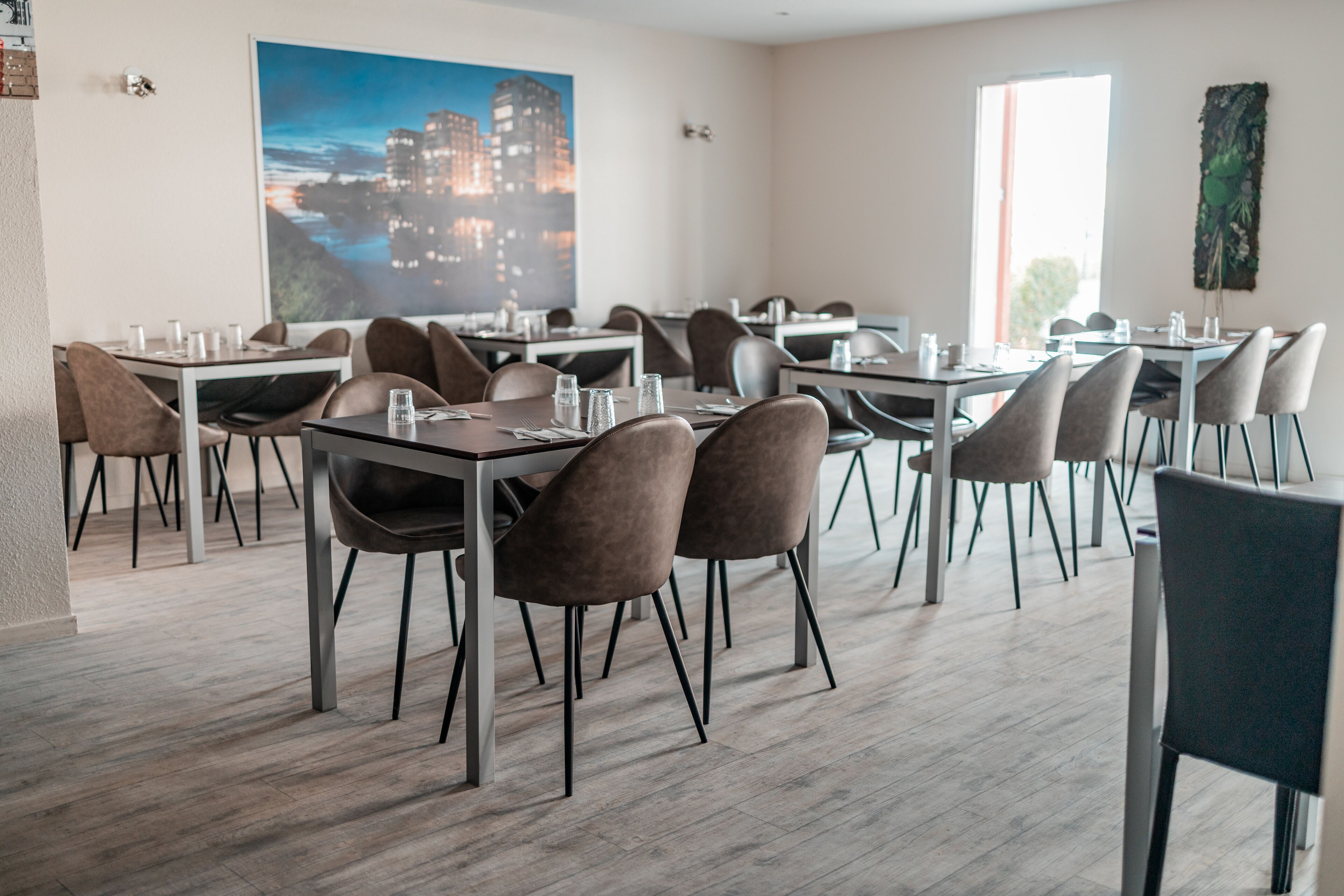 Un comedor moderno con varias mesas y sillas, un cuadro grande con una vista urbana en la pared, y un ventanal que proporciona luz natural.