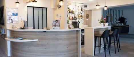 A modern hotel reception area with a wooden reception desk, bar area with bottles, and a seating area with chairs.