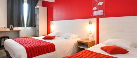 Une chambre d’hôtel avec deux lits, chacun recouvert d’une couverture rouge et agrémenté d’oreillers blancs. On y trouve un bureau avec une chaise et une petite table avec une lampe.