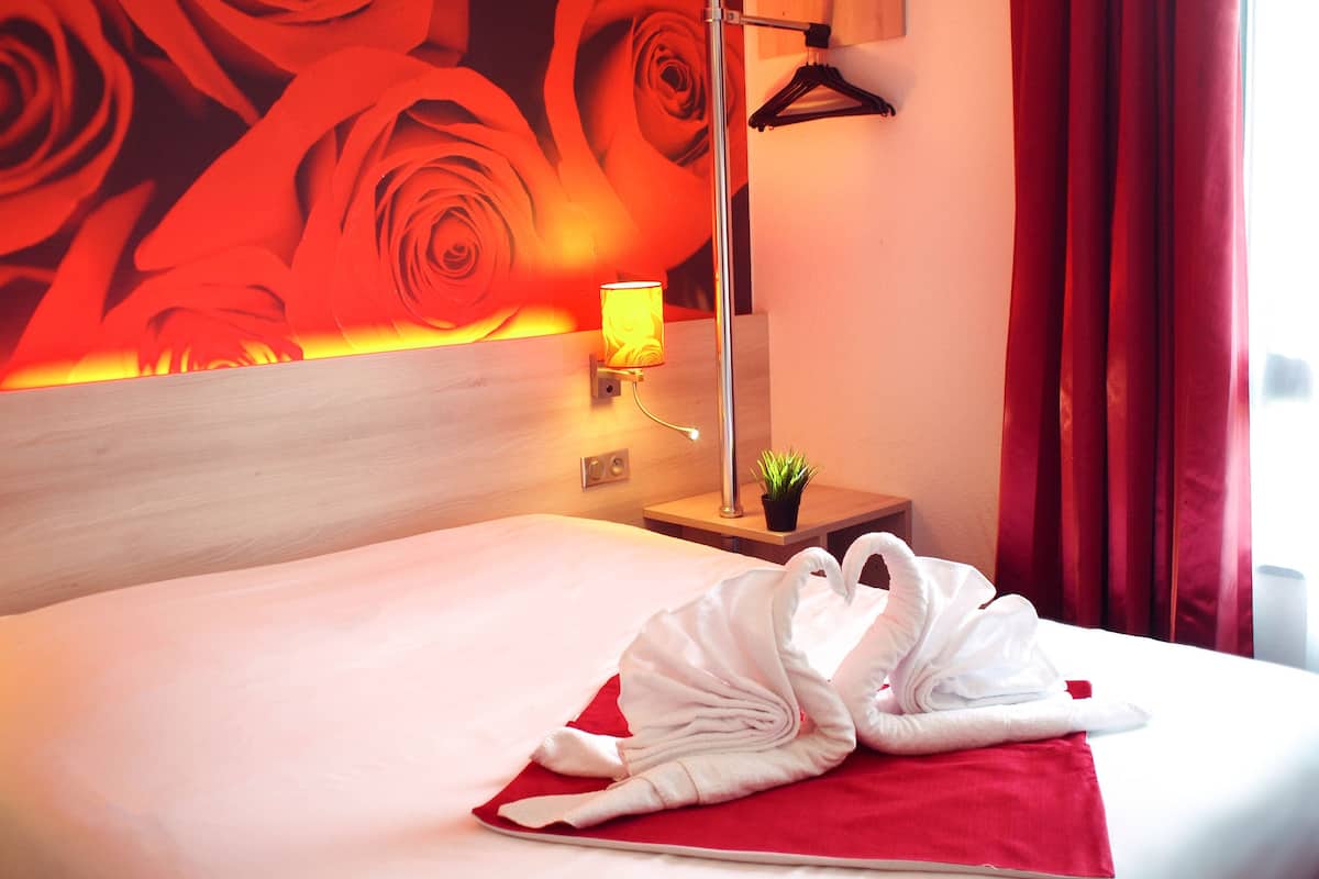A hotel room with a bed, a red pillow, a towel, a nightstand, a lamp, a small plant, and a red curtain.
