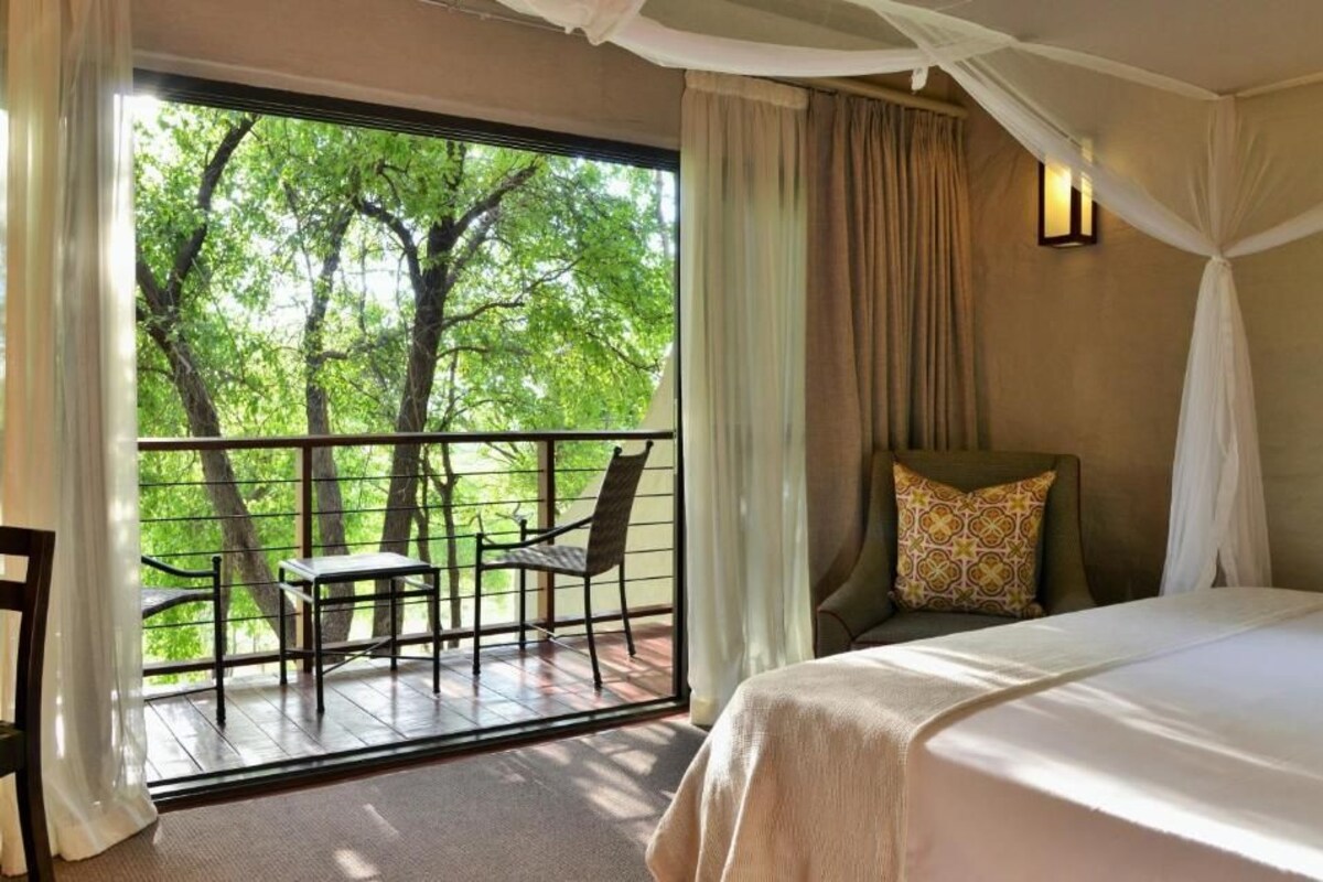 A room with a bed, a balcony with a table and chairs, and a view of trees.