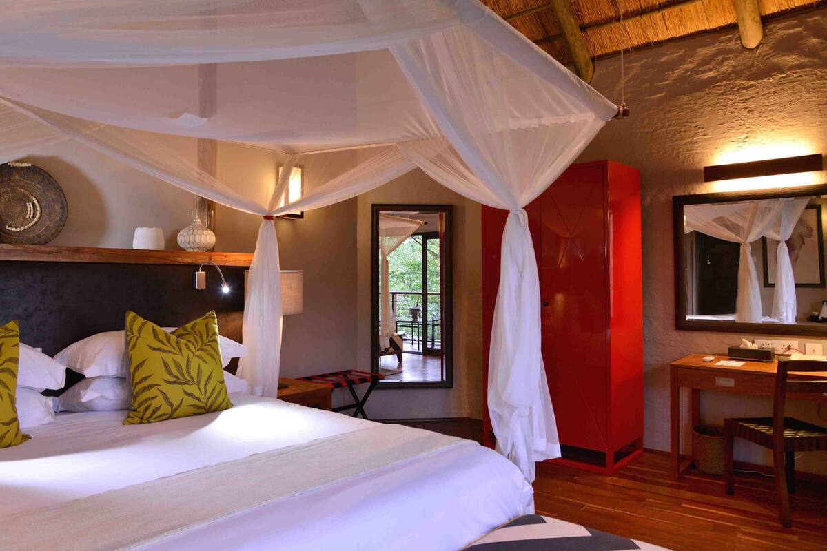 A bedroom with a canopy bed, a red cabinet, a mirror, and a window with a view of greenery.