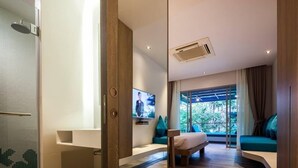 Een moderne hotelkamer met een houten vloer, een aan de muur bevestigde televisie, een groot raam met uitzicht op groen en een klein zithoekje met kussens.