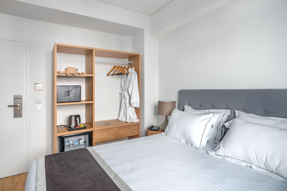A hotel room with a bed, a wardrobe, a bedside table with a lamp, and a small desk with a kettle and a phone.