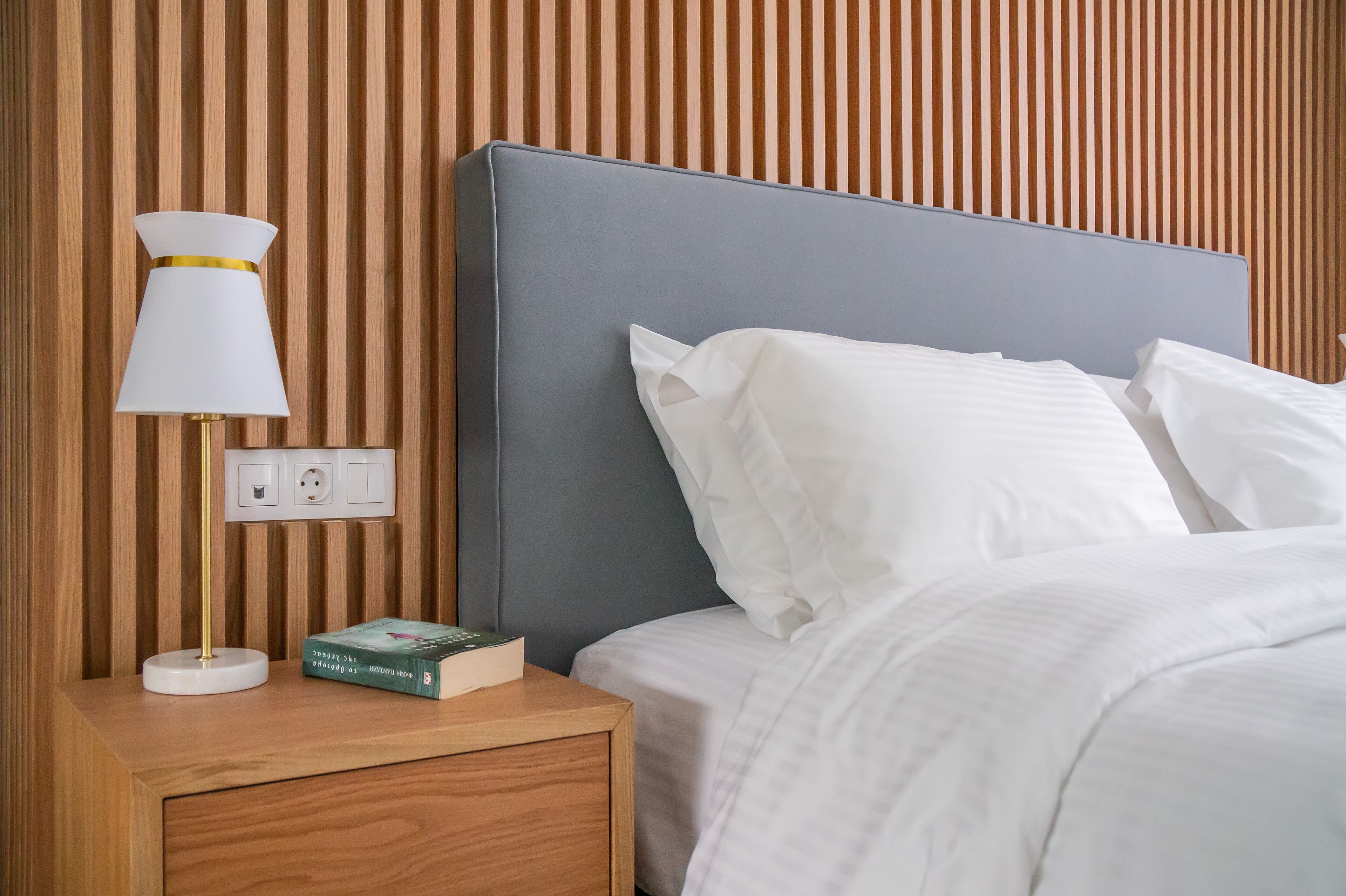 A neatly made bed with white linens, a bedside lamp with a white shade and gold trim, and a book on the nightstand.