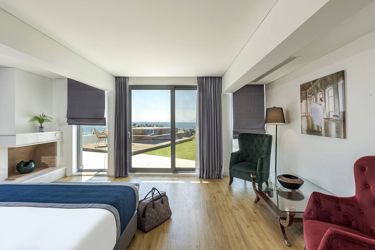 A modern hotel room with a large bed, a green armchair, a red armchair, a glass coffee table, and a view of the beach.