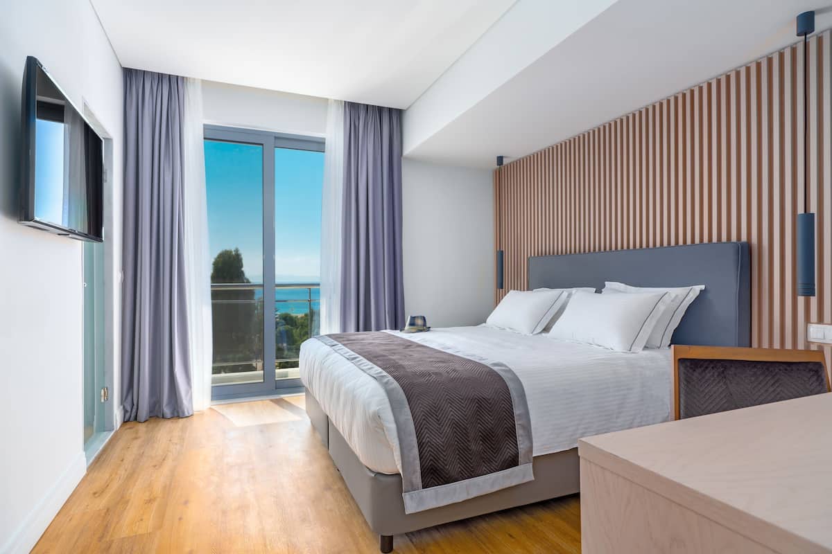 A modern hotel room with a large bed, a TV, a wooden panel wall, and a balcony with a view.